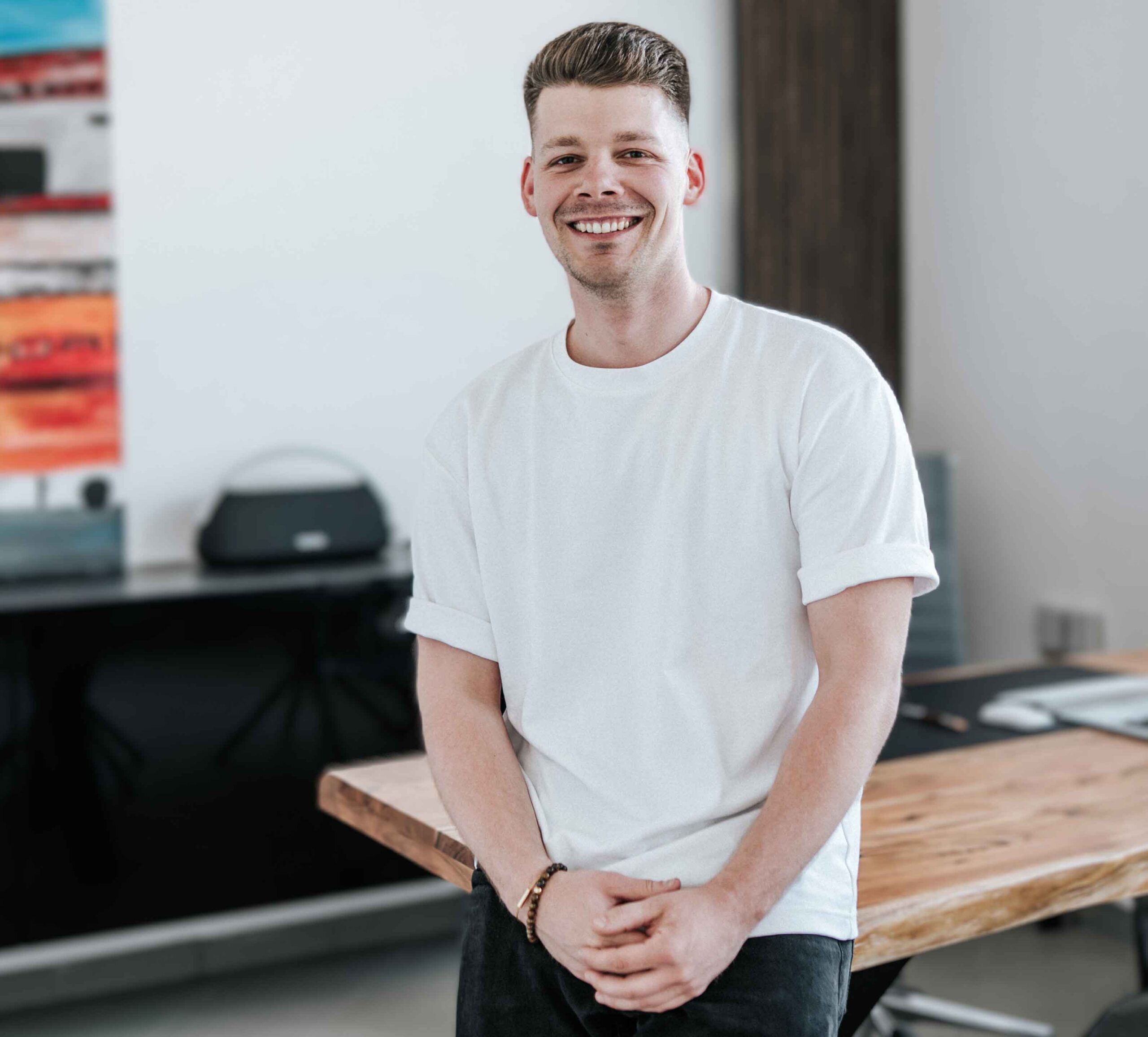 Premiumfahrzeuge kaufen & verkaufen 4 Philip Pelzer Premium Autohändler Junger Mann in weißem T-Shirt steht vor einem Holztisch in einem modernen Büro mit Laptop und abstraktem Gemälde