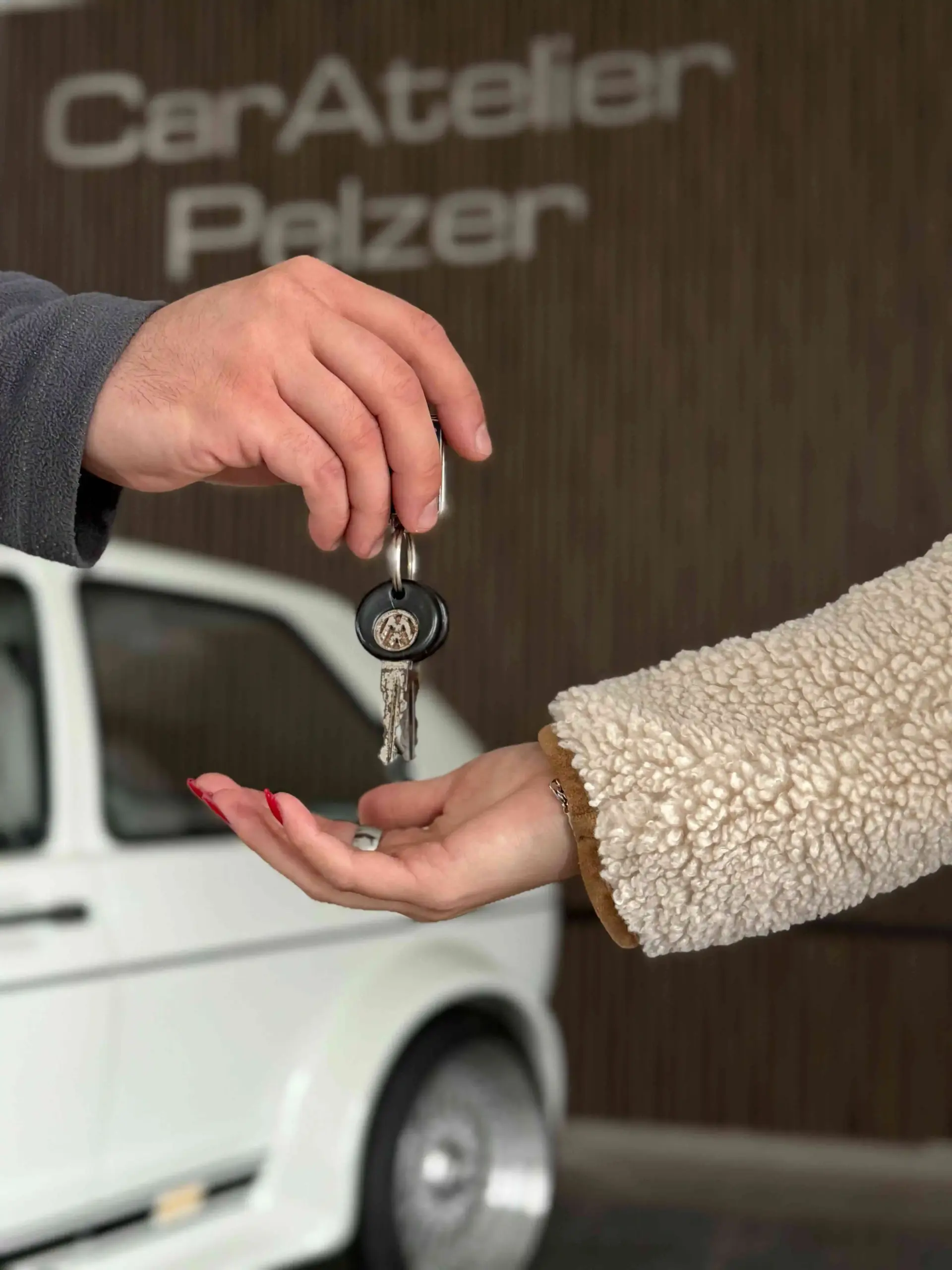 Premiumfahrzeuge kaufen & verkaufen 2 Hand übergibt Autoschlüssel an andere Hand vor Auto und Schild 'CarAtelier Pelzer'